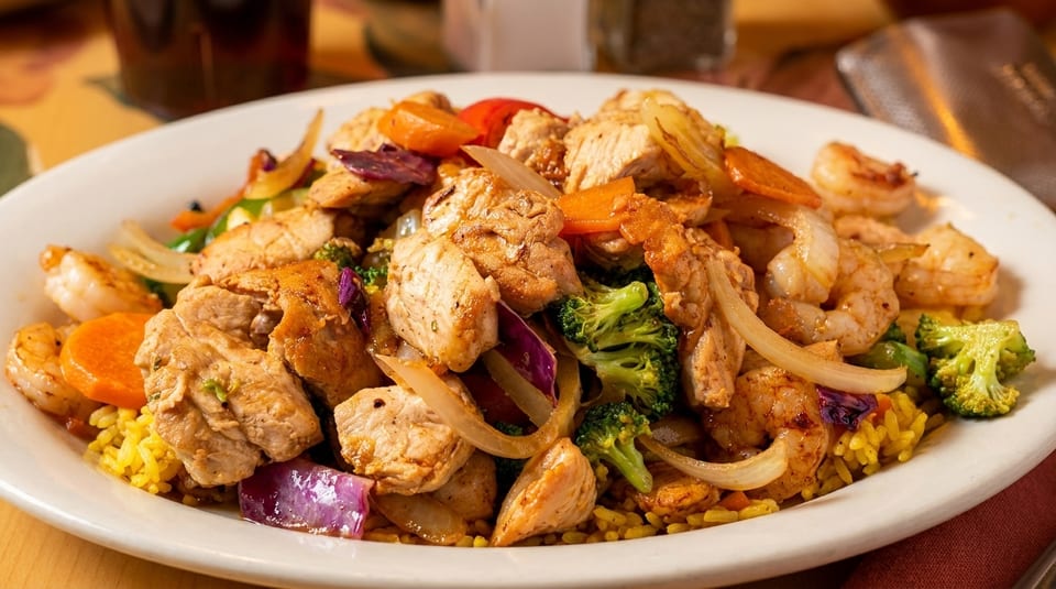 A white plate filled with chicken and shrimp stir-fry over yellow rice, with vegetables.