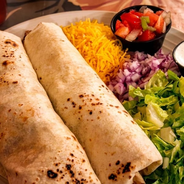 Two burritos with shredded cheese, lettuce, diced onions, pico de gallo, and sour cream.