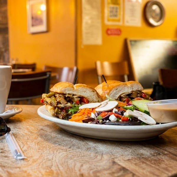 A sandwich and salad plate with a coffee cup and sunglasses on a rustic wooden table.